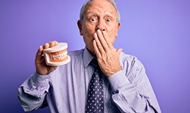 Older man covering his mouth with one hand while holding artificial teeth in the other in front of a purple background